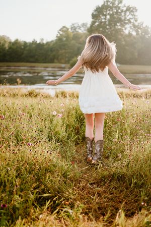 Person in white dress and cowboy boots dances through a wildflower meadow at golden hour near a lake.