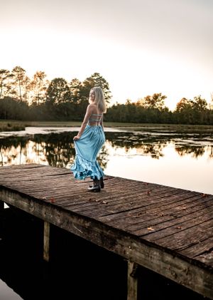 Scenic lakeside senior photo on wooden dock with flowing blue dress during sunset.