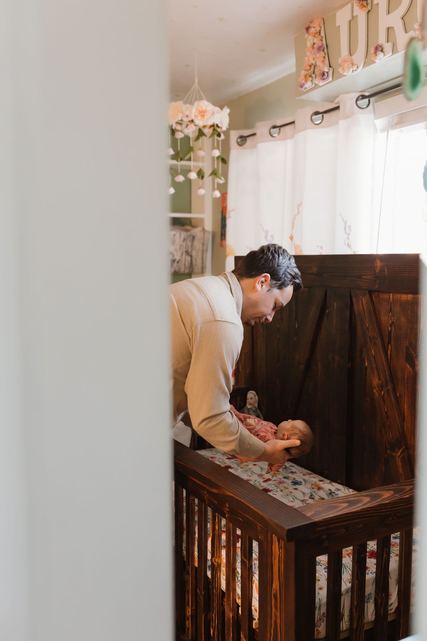 A parent stands over a wooden crib in a rustic nursery decorated with floral accents and sheer curtains. Colorado Springs Photographer.