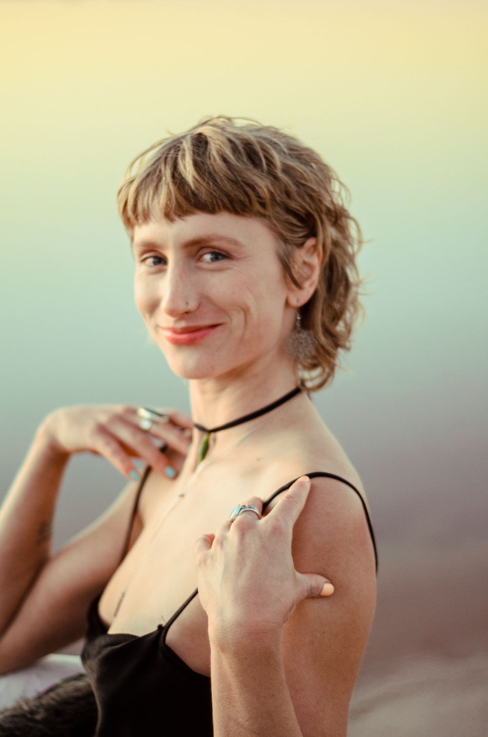 Portrait in warm lighting showing jewelry and a black choker necklace against a soft gradient background.