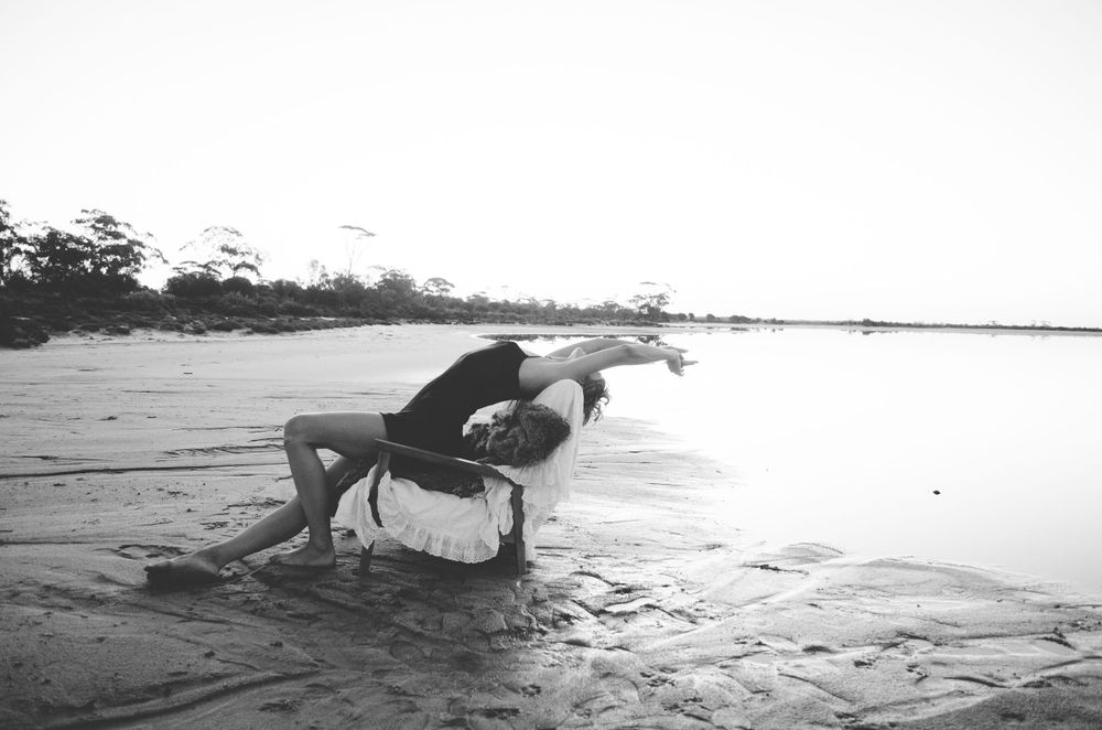 Artistic black and white photo of a dancer posed gracefully on a chair at the beach during sunset.