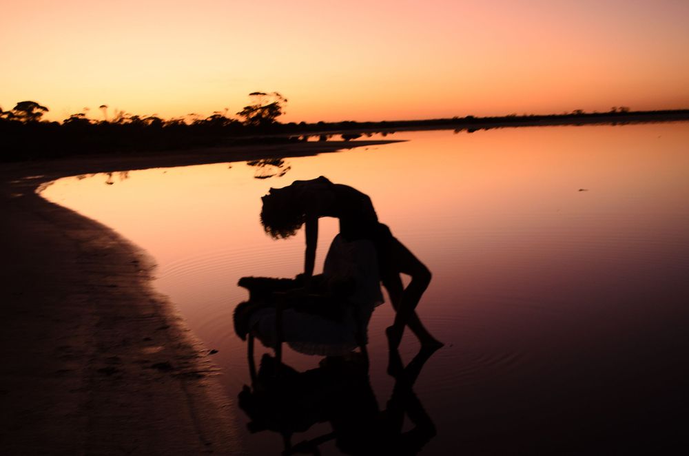 Silhouetted figure performing an expressive dance pose against a pink sunset over calm waters.