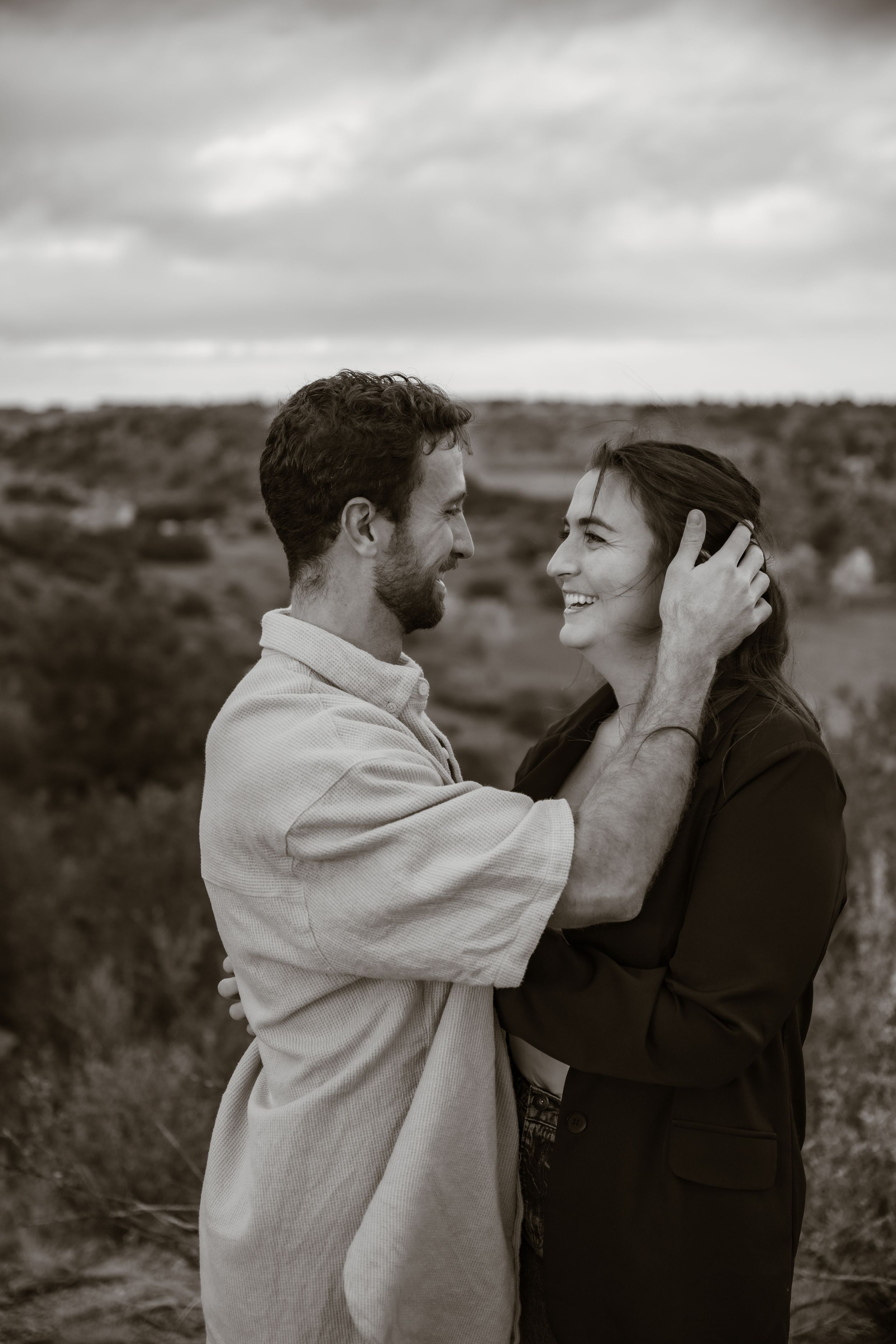 A couple shares an emotional moment against a cloudy sky in black and white photography.