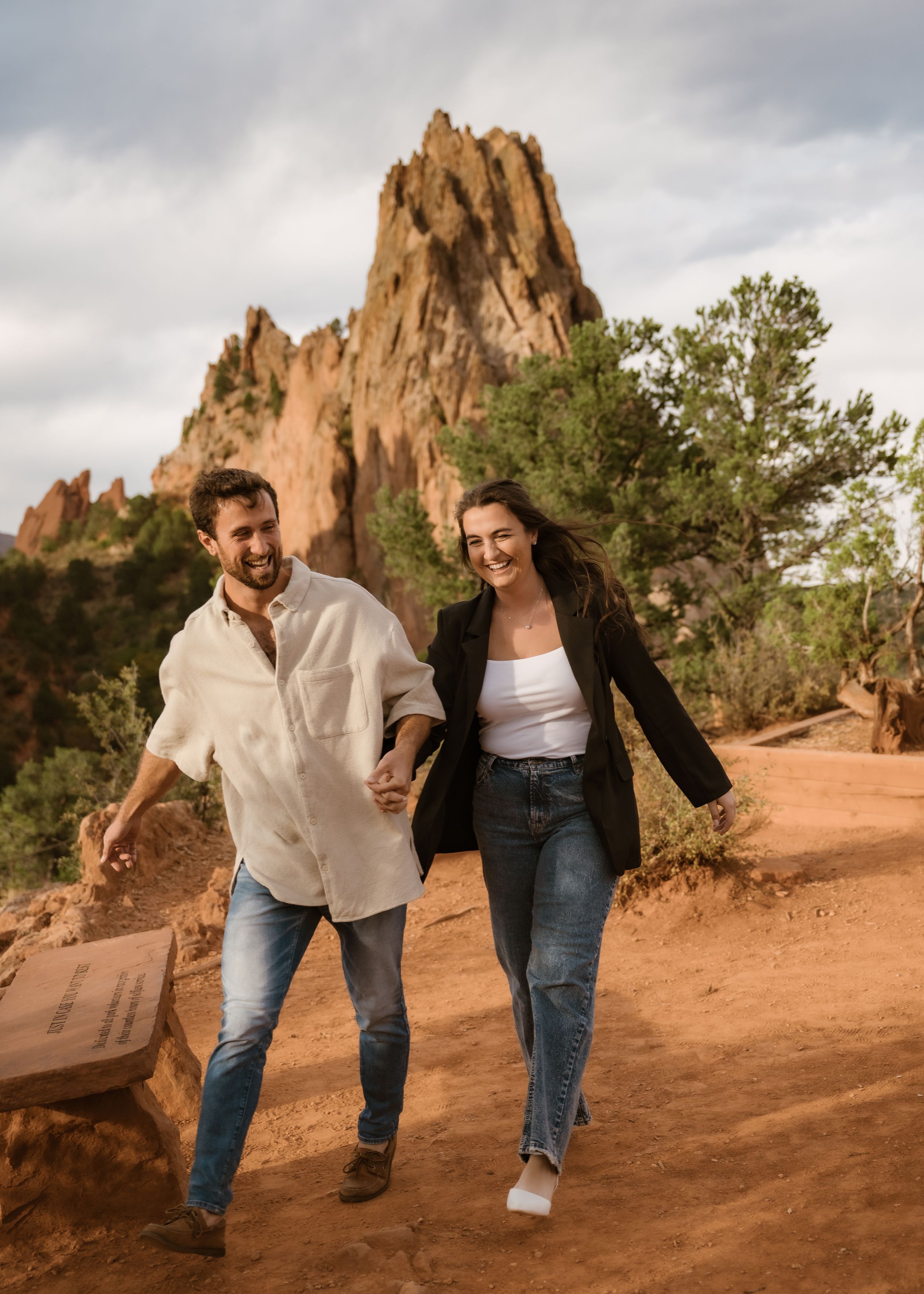 A playful couple walks together on red desert rocks with towering formations in the background.