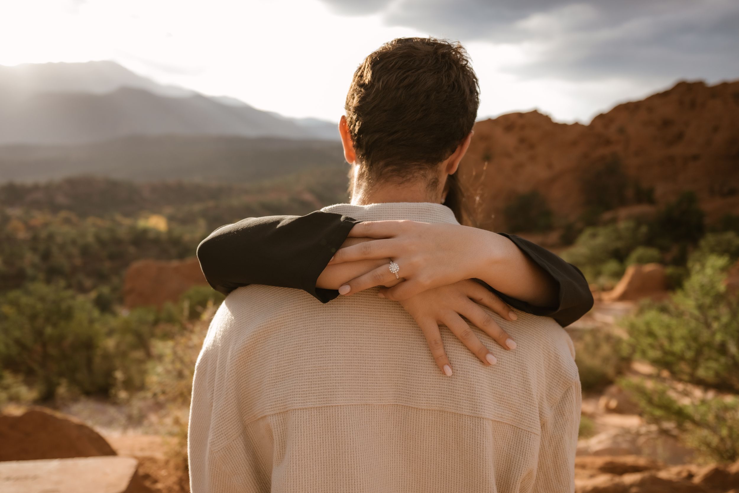 A romantic couple embraces against a backdrop of red rock formations and desert landscape at sunset.