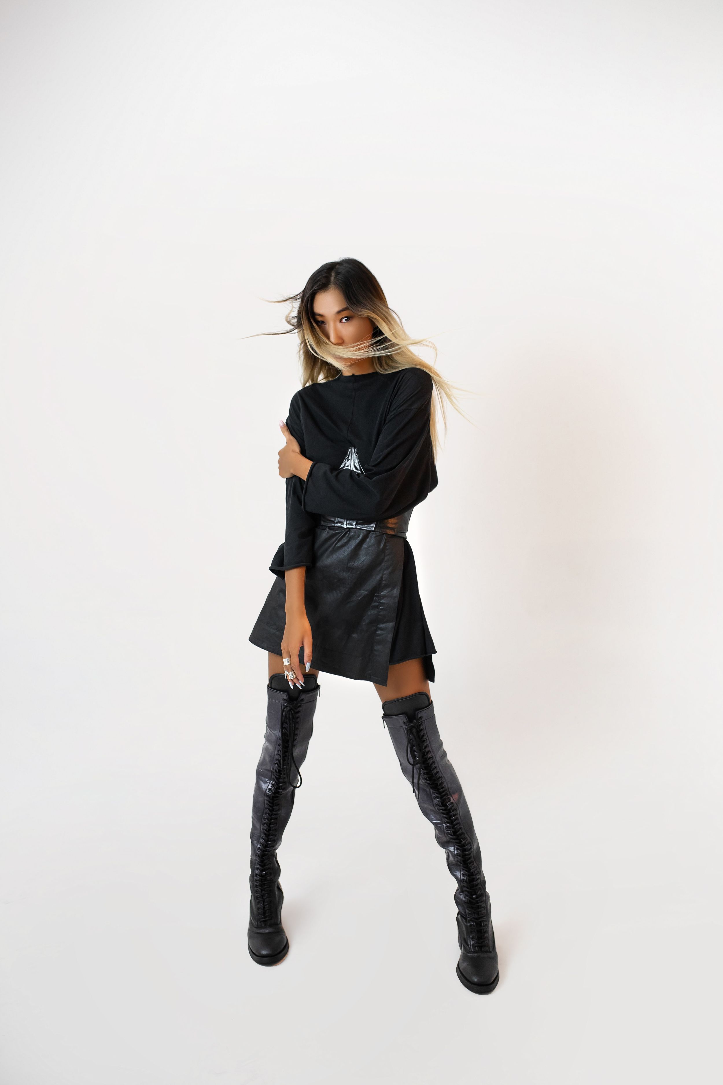 Fashion model in black outfit and thigh-high boots poses against white background.