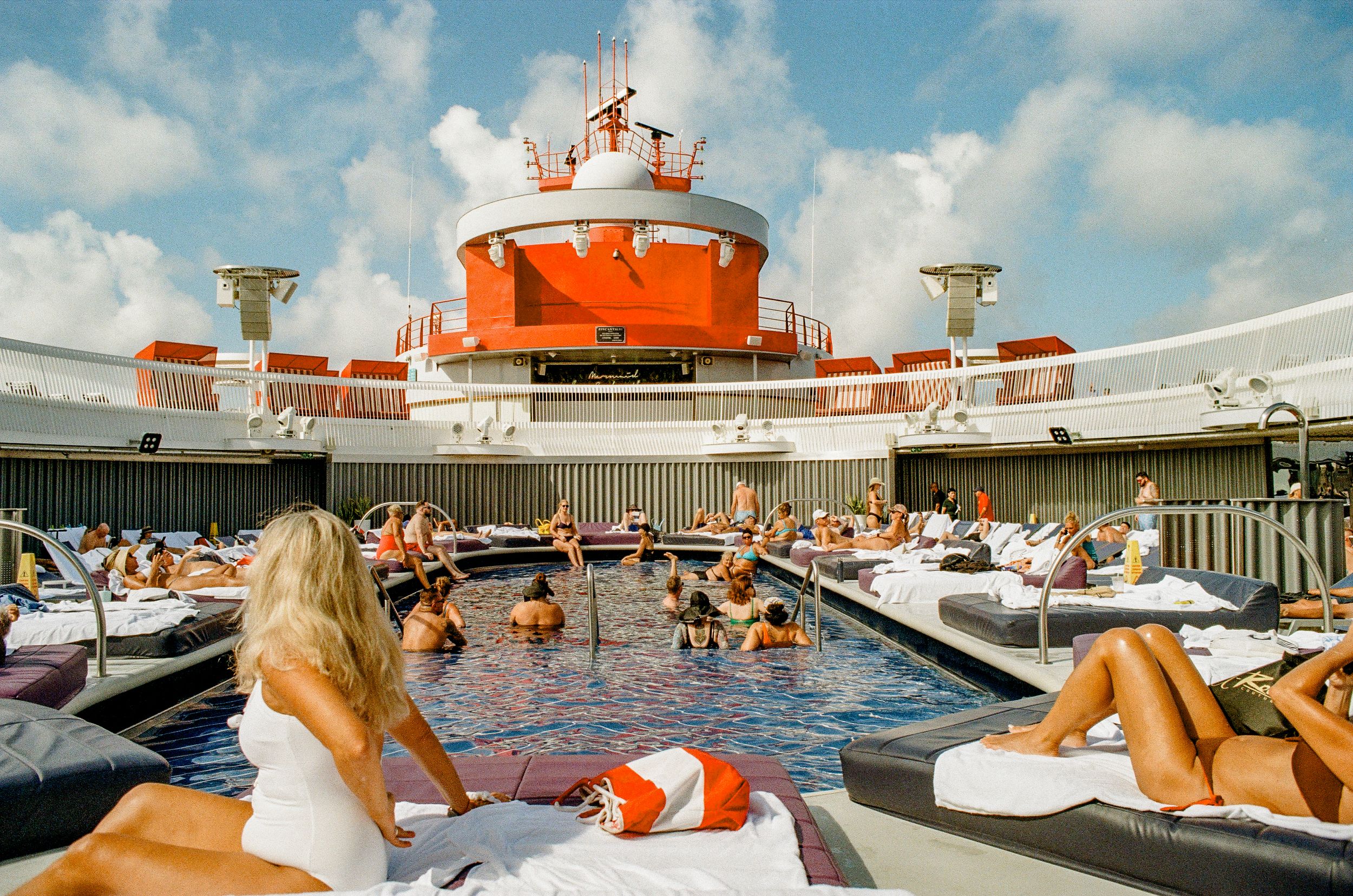 Crowded cruise ship pool deck with orange and white color scheme and passengers lounging in sunlight.