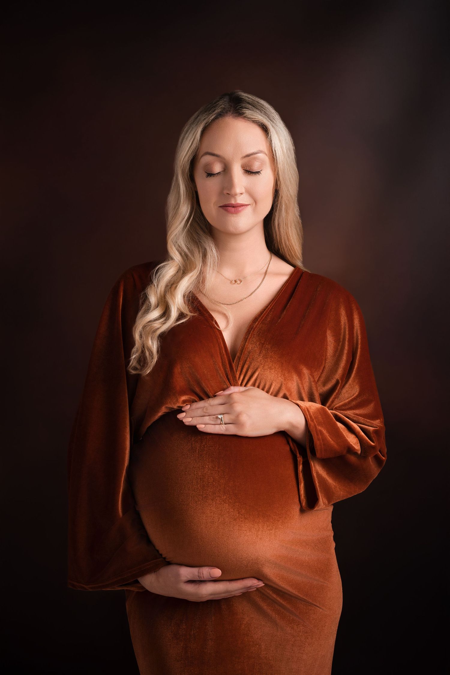 Elegant maternity portrait in copper velvet dress against dark background.