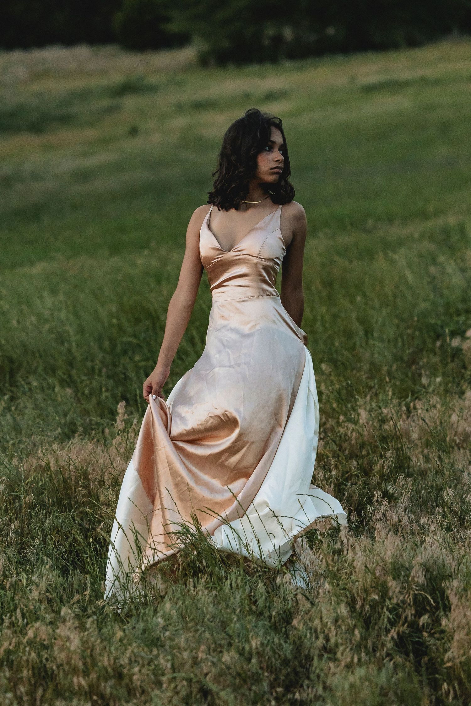 A figure in a flowing cream dress sits gracefully in a sunlit meadow of tall grass.