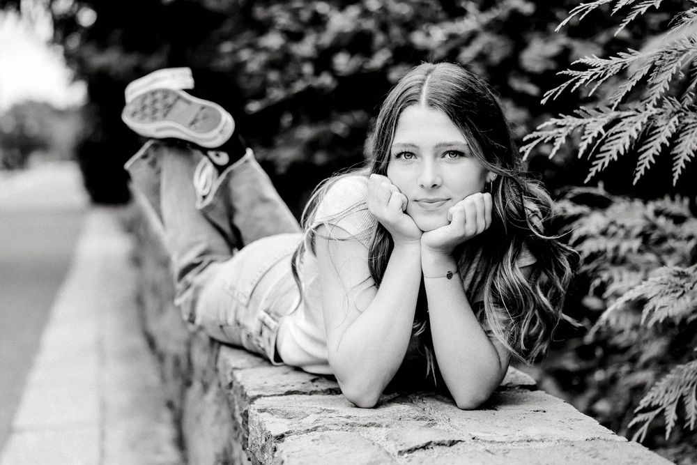 Black and white photo of a teen girl lying on a stone wall surrounded by greenery.