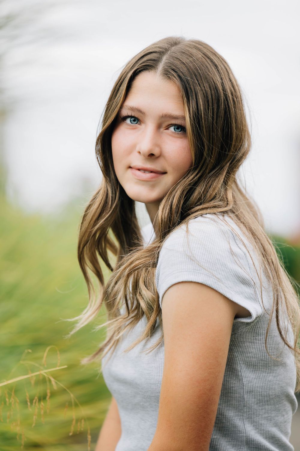 Teen girl with long wavy brown hair in a gray tee smiling outdoors in nature.