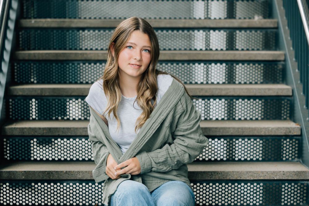 Young woman in green hoodie and jeans sits on metal stairs smiling softly in a senior portrait photo.