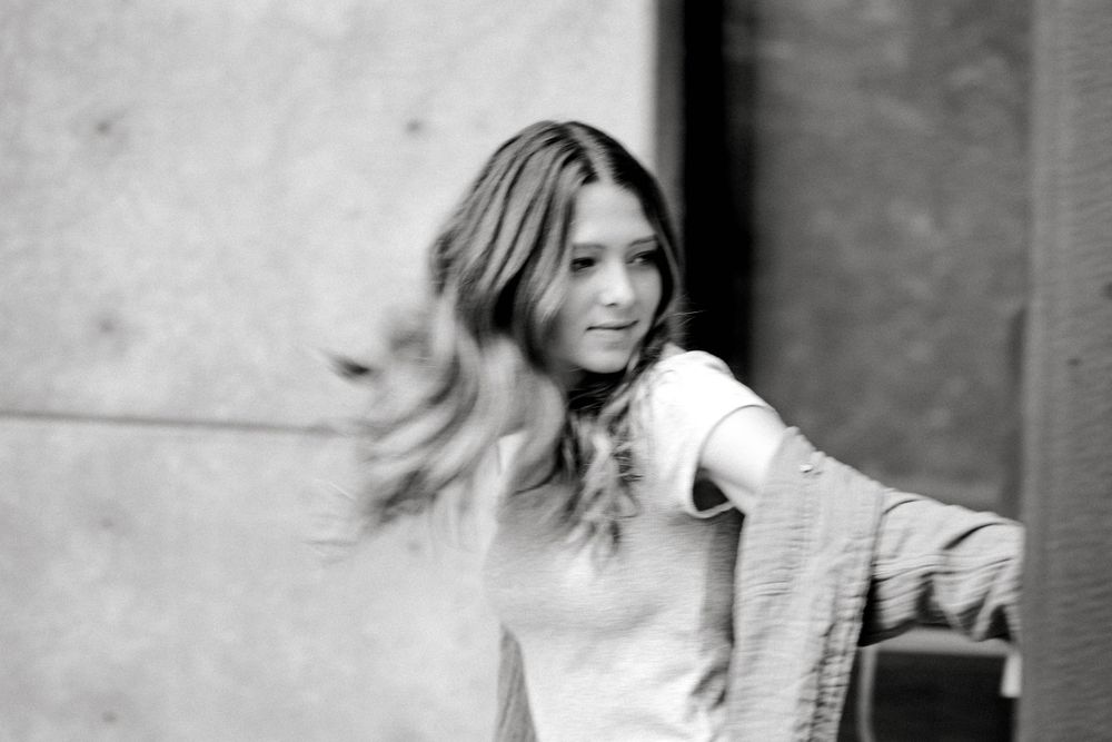 Black and white photo of a young woman with flowing hair wearing a light top and open jacket outdoors.