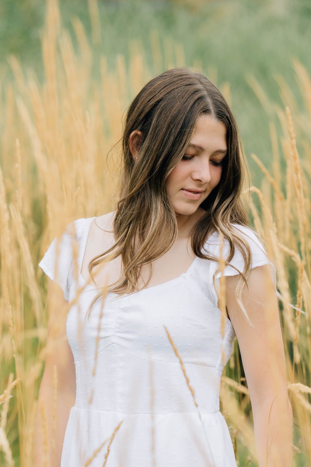 Young brunette woman in white dress standing in golden tall grass field looking down peacefully.