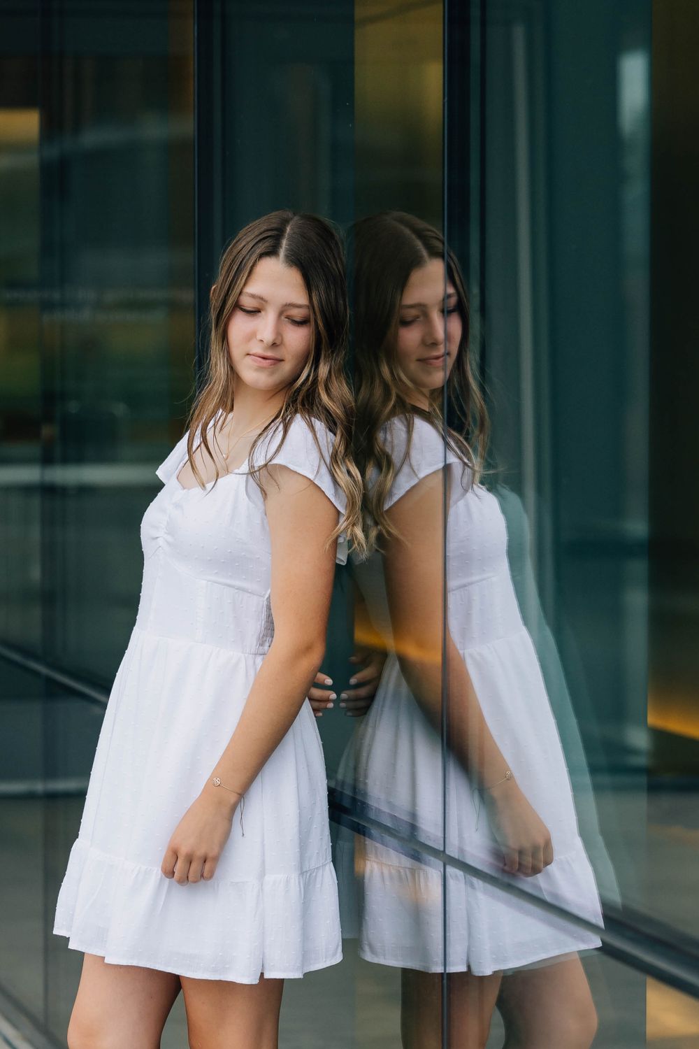 Teen girl in white dress leaning against glass wall with reflection visible in urban setting.