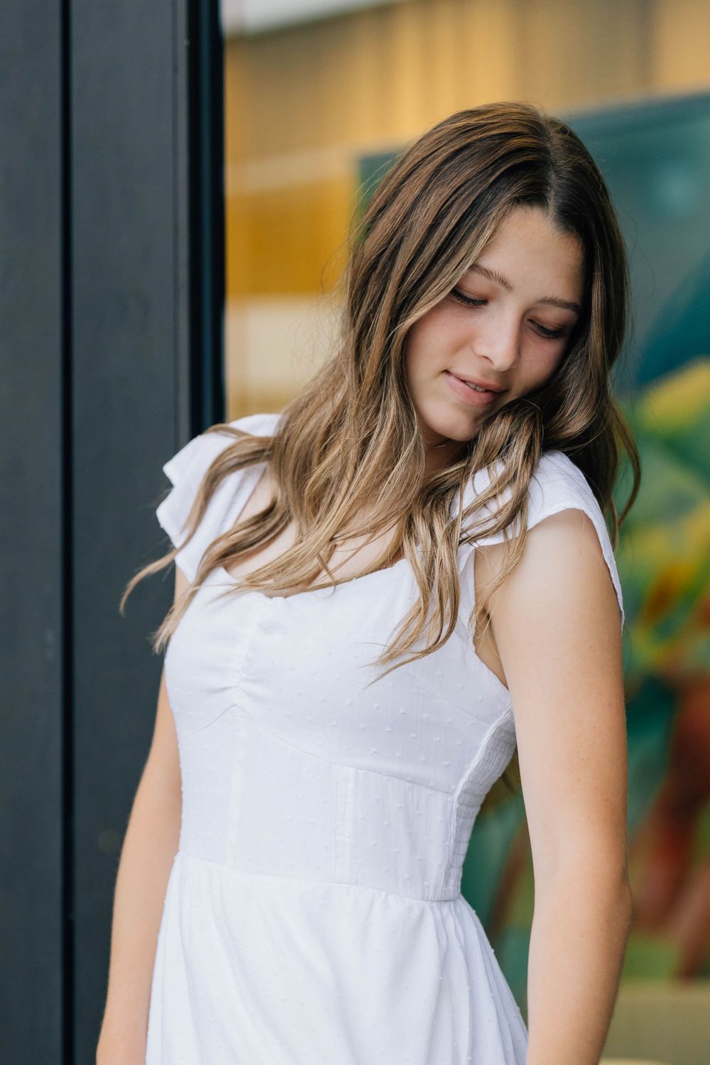 Young woman with wavy highlighted hair in white dress looking down near colorful wall backdrop.