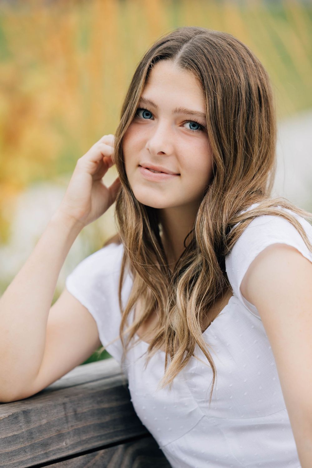 Teen girl with blue eyes and long wavy hair in white tee smiling outdoors near wooden surface.