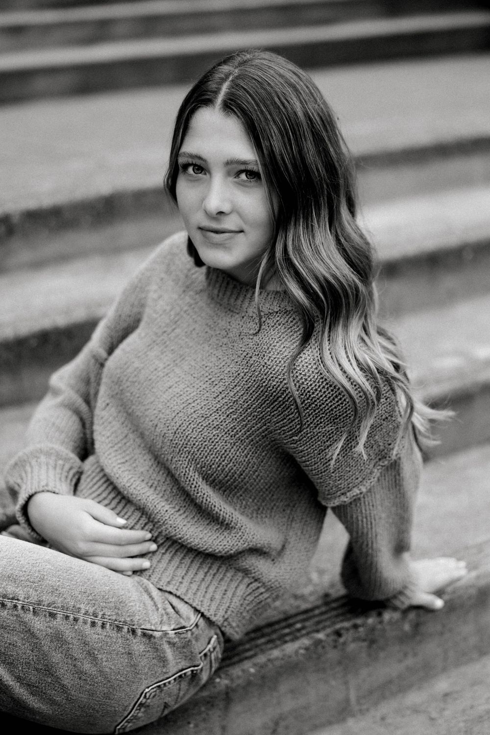 Black and white senior portrait of young woman in knit sweater and jeans sitting on stone steps.