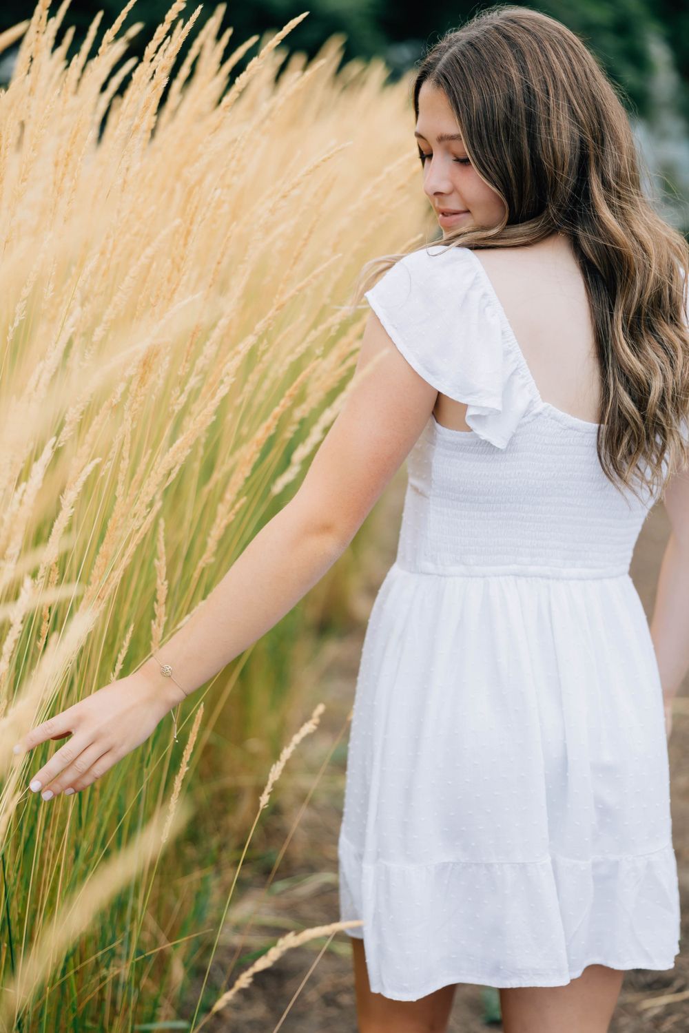 Young woman in white flutter-sleeve dress reaching out to touch tall golden grass in a field.