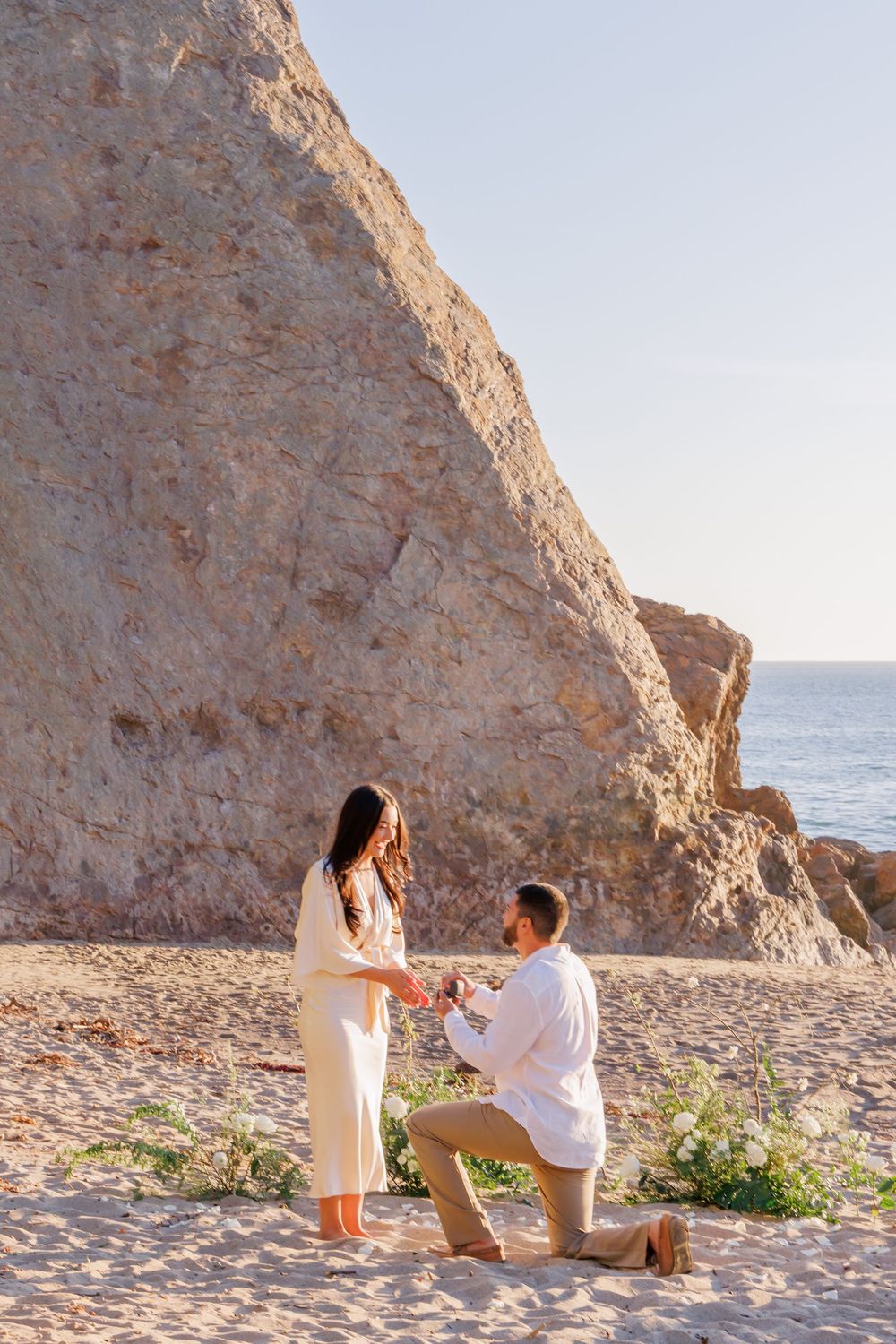 Man proposing on one knee to woman on sandy beach at sunset with dramatic rocky cliffs in background.