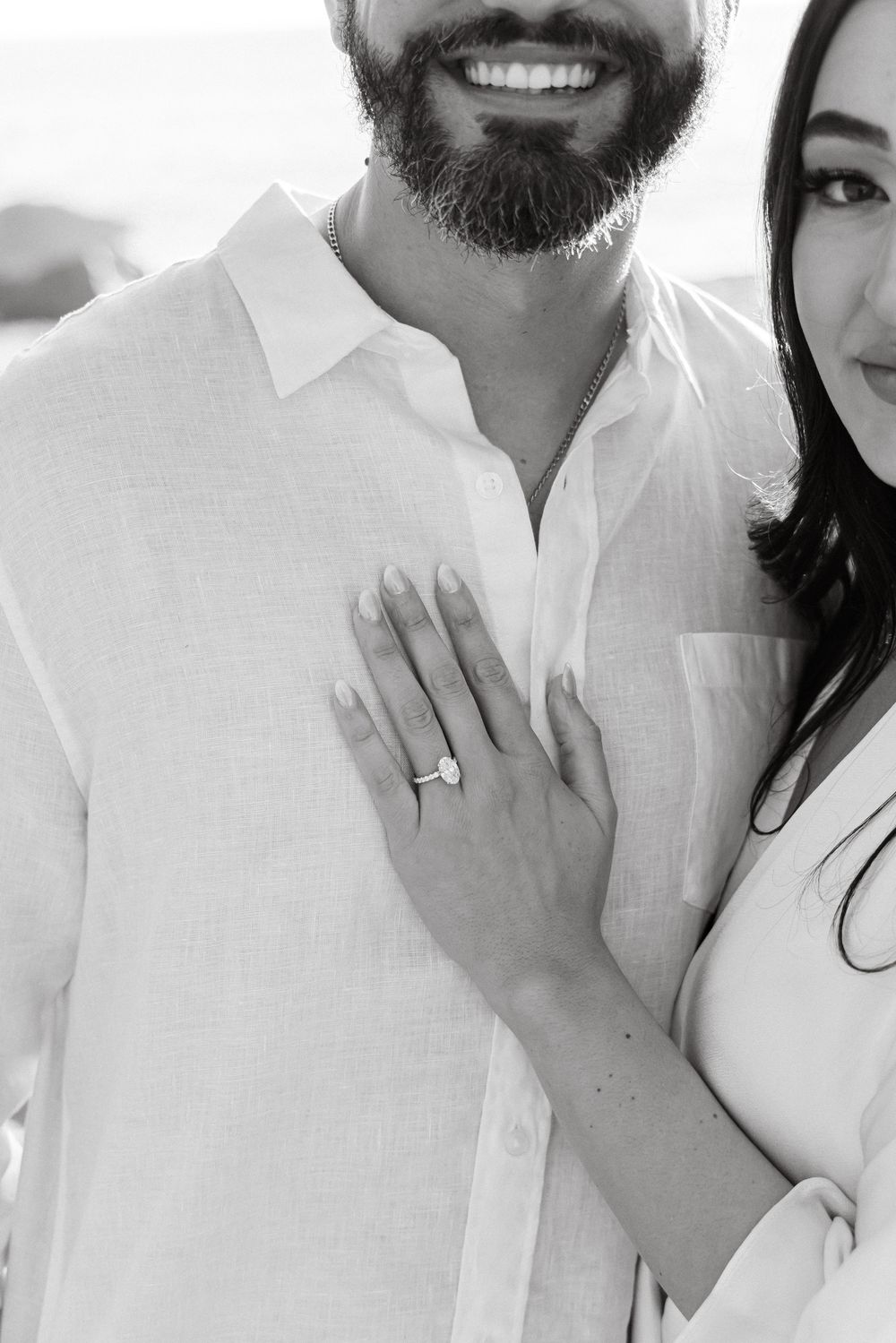 Black and white close-up of smiling bearded man with woman showing diamond engagement ring on hand.