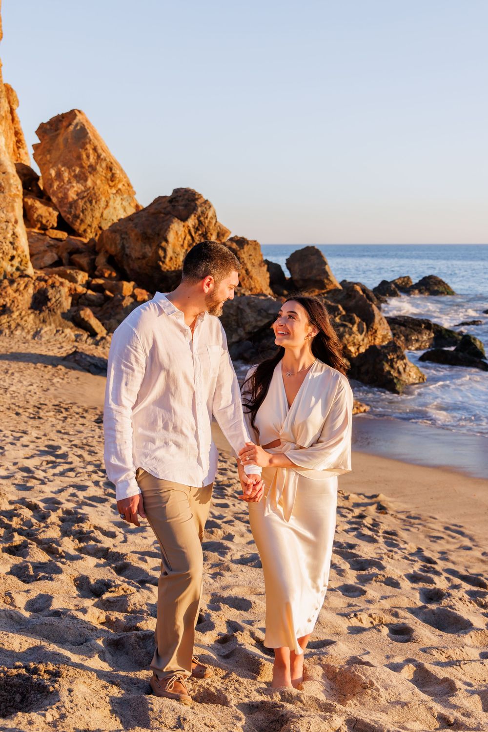 Happy engaged couple holding hands walking on sunny beach with rocky coastline in background.