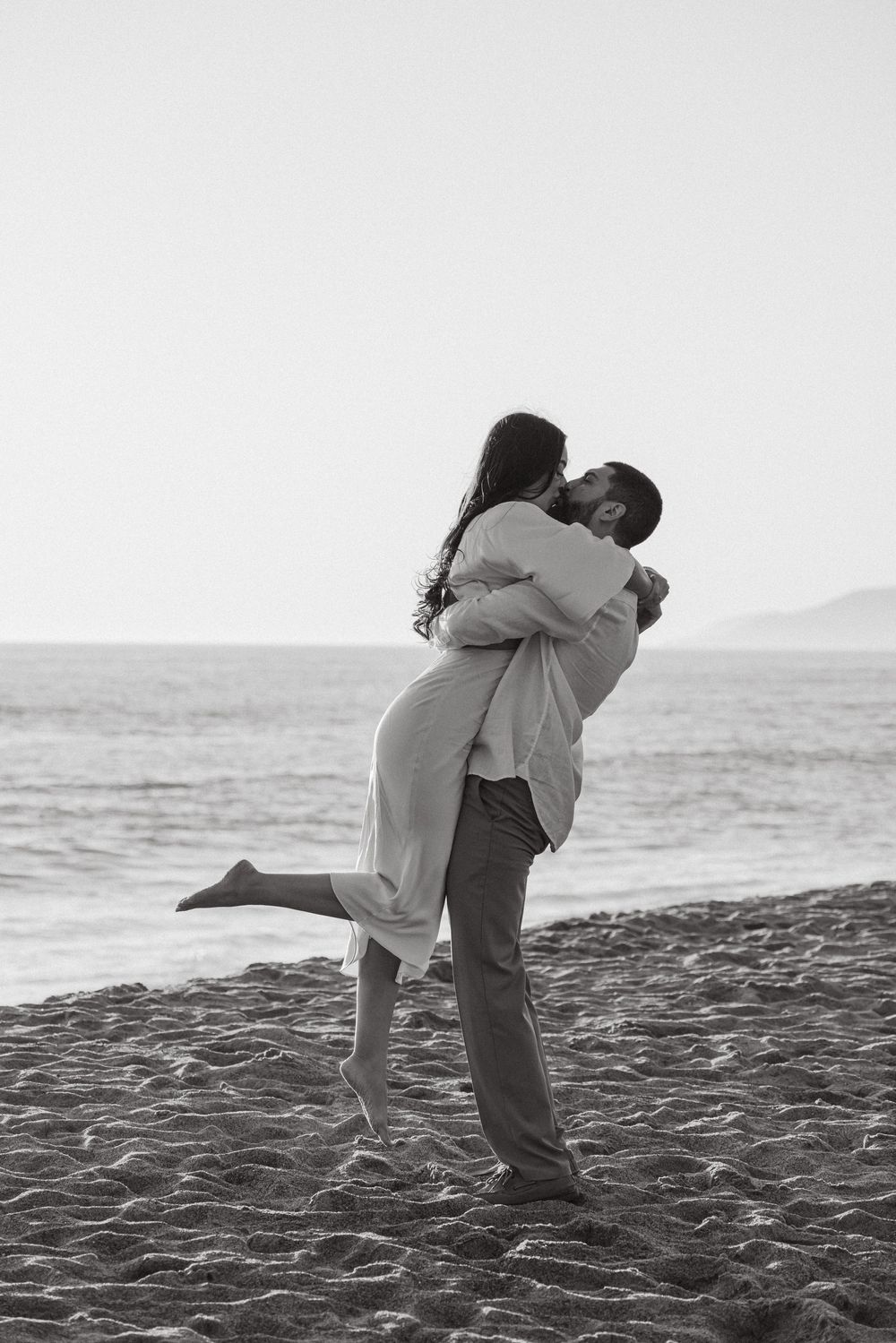 Black and white romantic photo of man lifting woman in joyful embrace on scenic ocean beach.