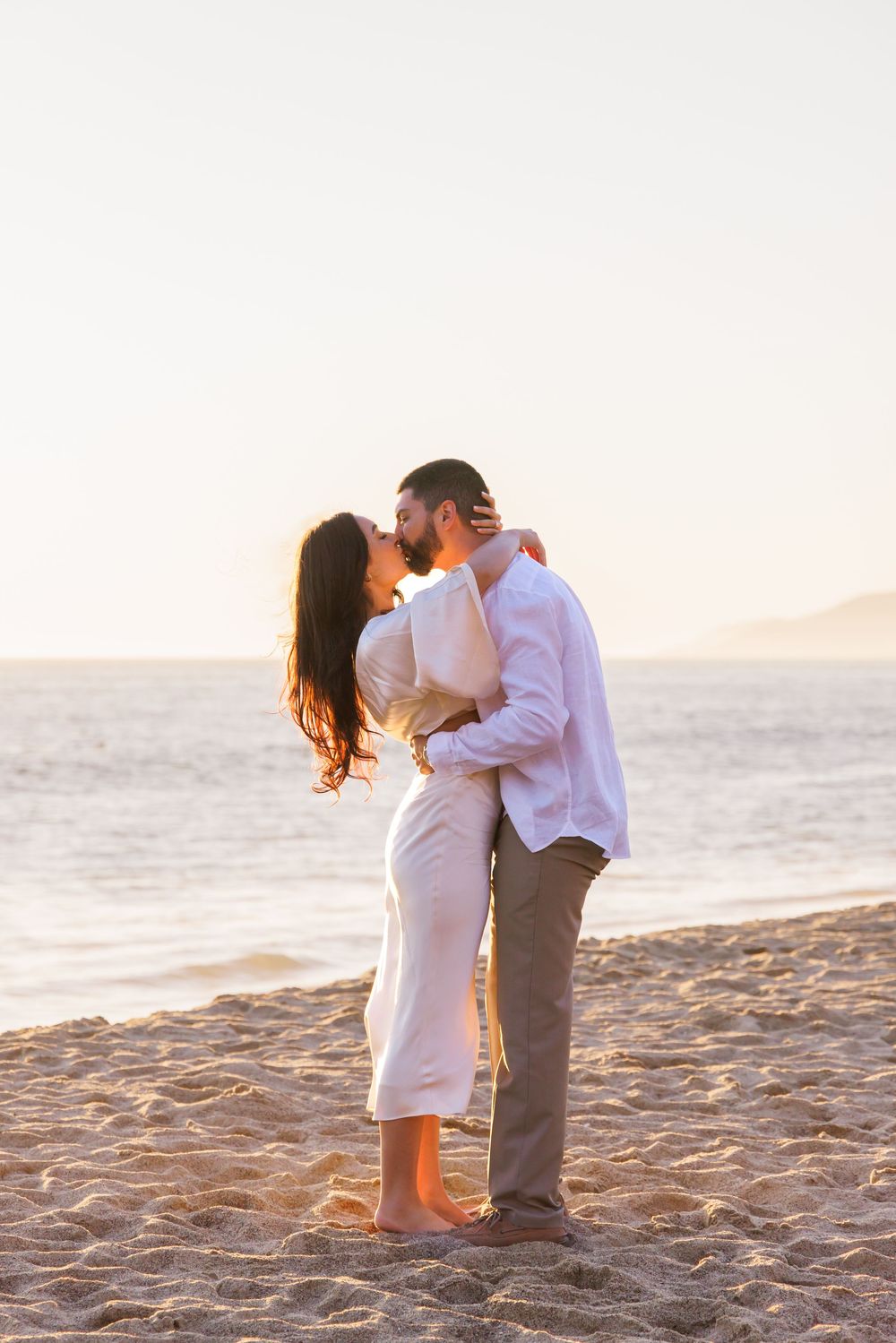 Couple kissing barefoot on sandy beach at sunset, woman in white dress, man in linen shirt.