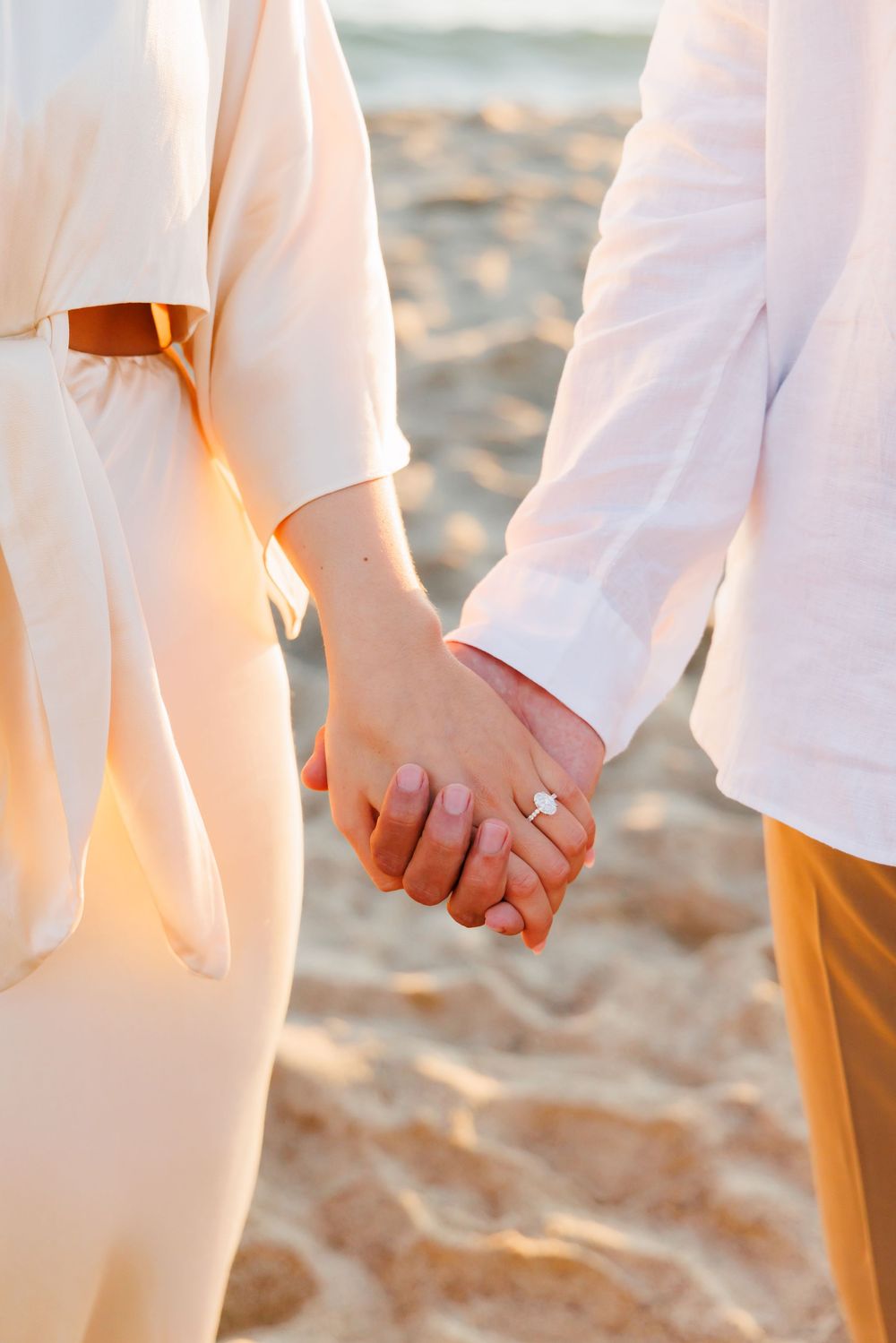 Close-up of engaged couple holding hands on sandy beach showing diamond engagement ring at sunset.