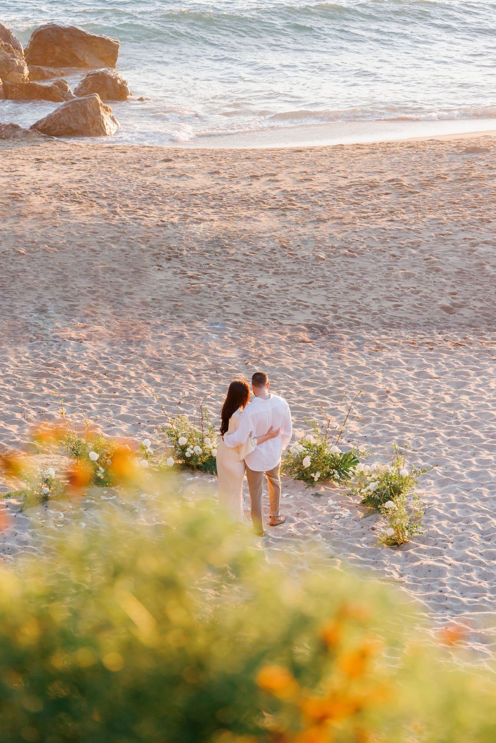 Couple embracing on sandy beach surrounded by floral arrangement with ocean and rocks behind them.