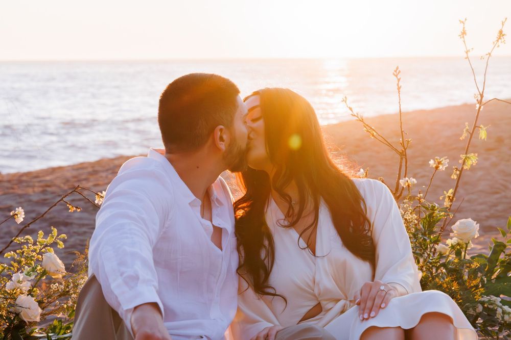 Couple sharing romantic kiss while sitting among white flowers on beach during golden sunset glow.