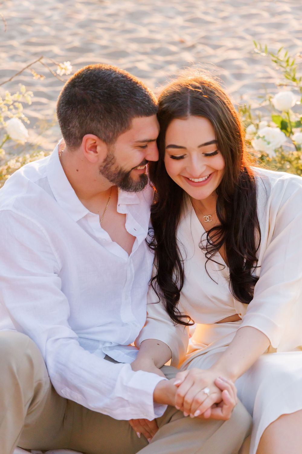 Newly engaged couple sitting on beach at sunset smiling and holding hands with white flowers nearby.