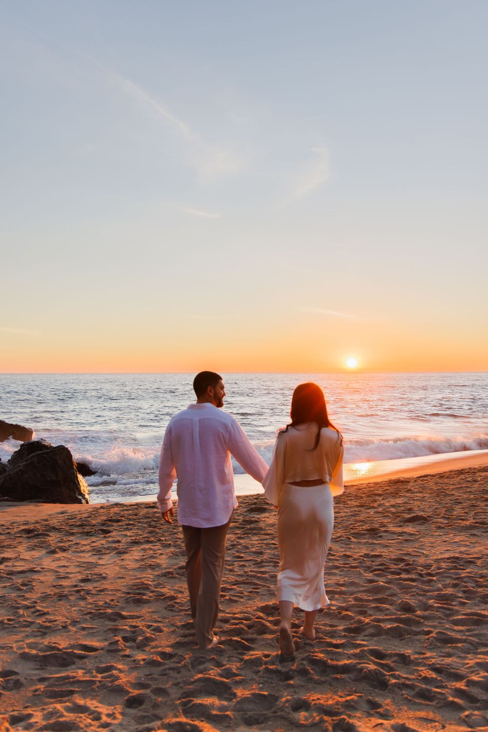 Couple holding hands walking on sandy beach toward stunning orange sunset over calm ocean waves.