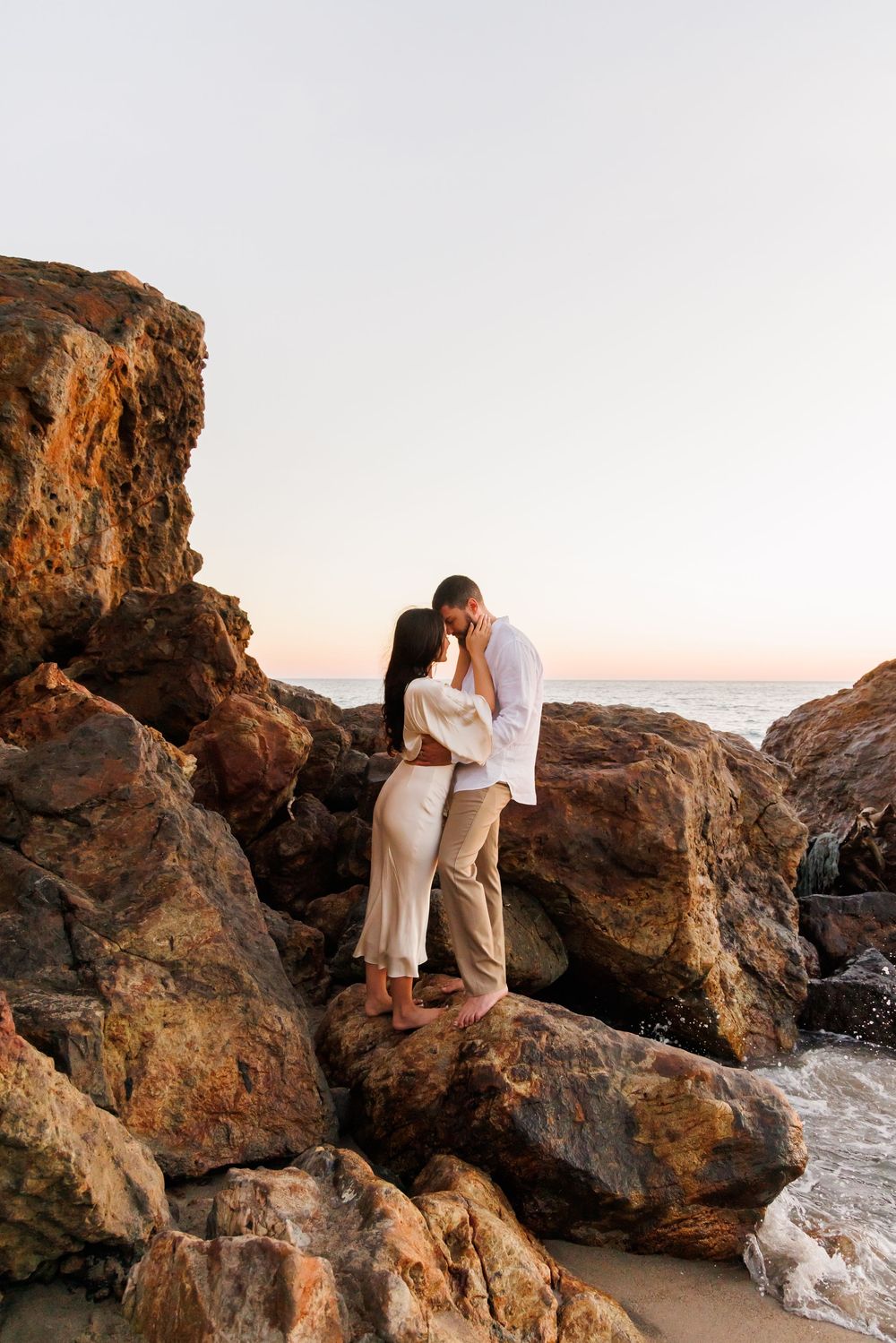 Romantic couple embracing and kissing on rocky coastal boulders during golden hour sunset at beach.