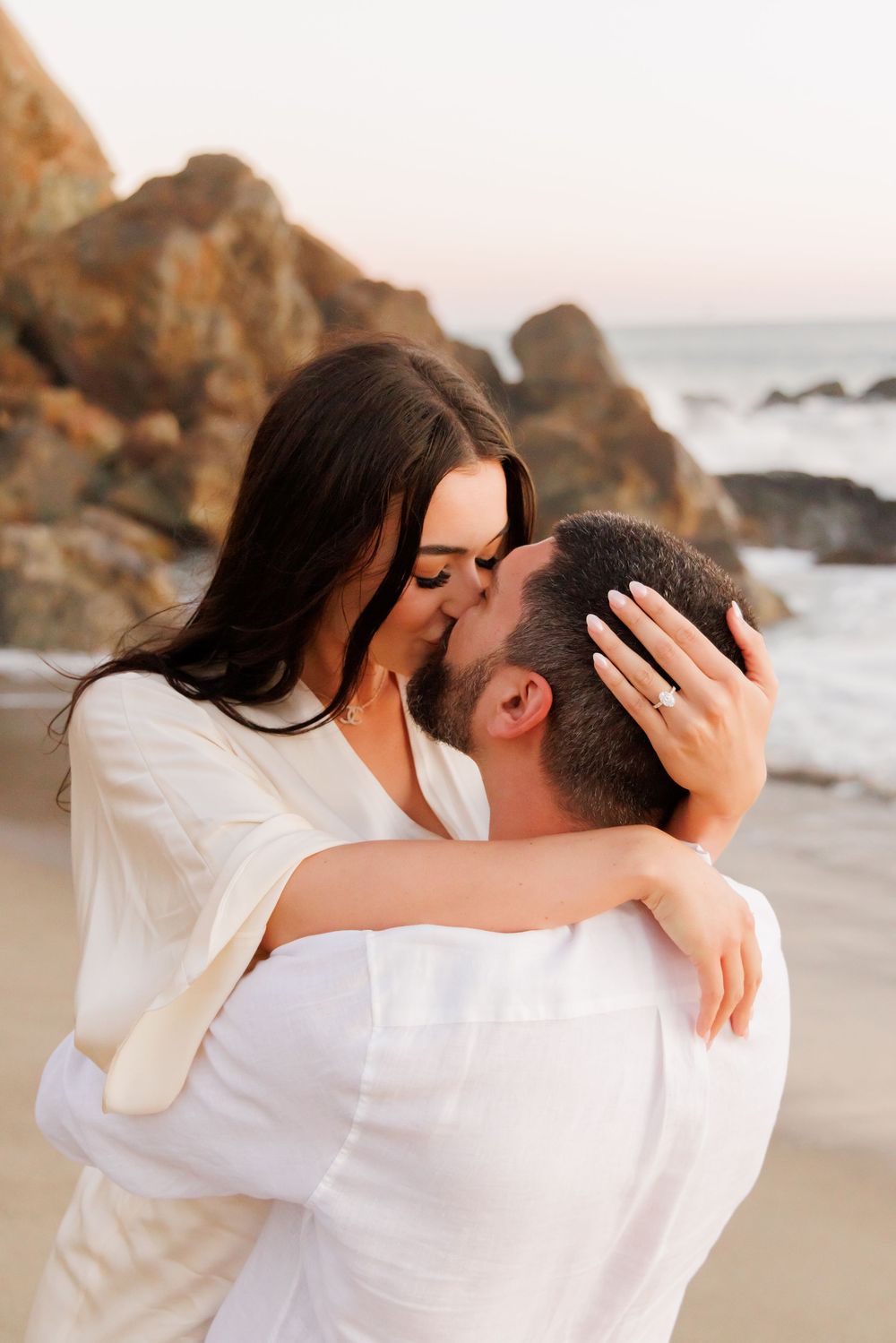 Engaged couple sharing passionate kiss on beach, woman displaying diamond engagement ring on hand.