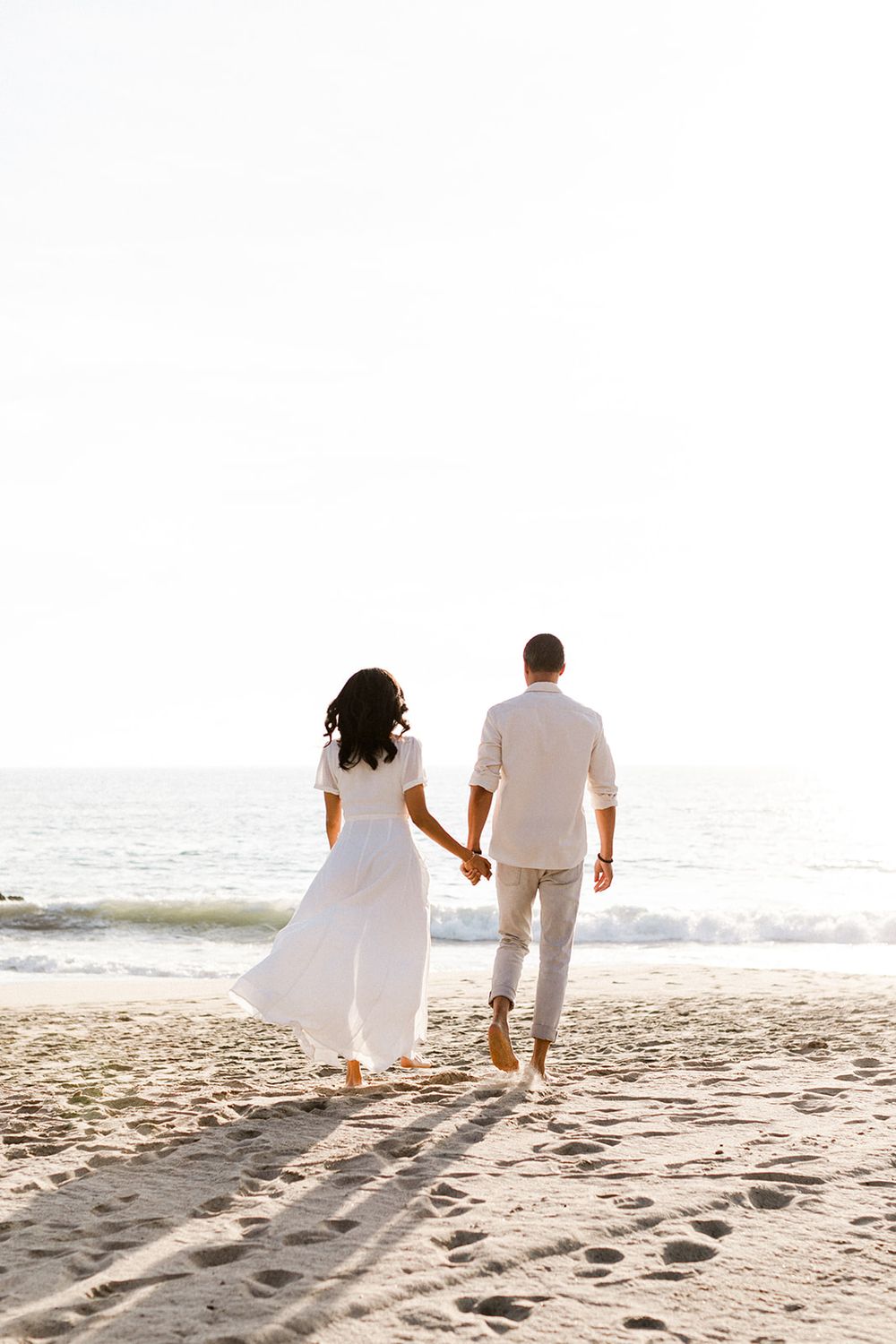 Romantic couple holding hands walking barefoot toward ocean waves on sandy beach in white outfits.