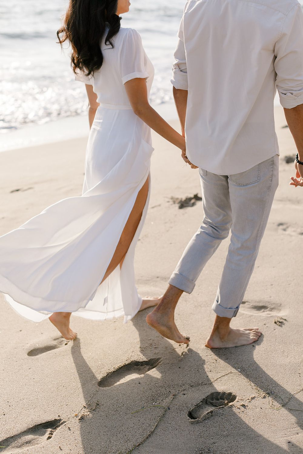 Close-up of barefoot couple holding hands walking on sandy beach leaving footprints, white flowing dress.