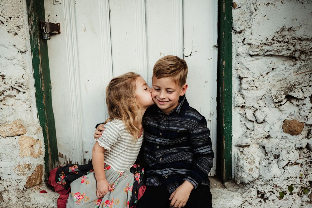 A couple embraces and laughs together against a rustic white stone wall with green trim.