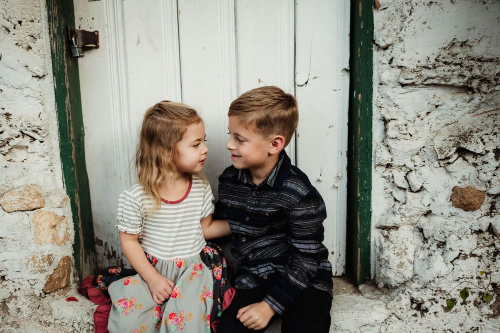 Children share playful interactions against a rustic white stone wall with green trim during an outdoor photo session.