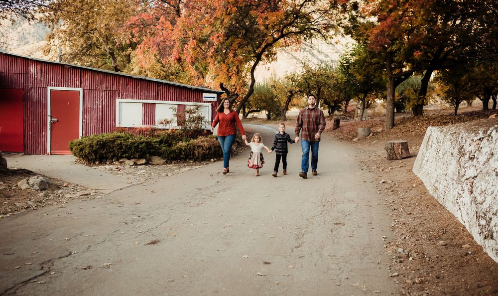 A family walks together on a dirt path surrounded by autumn trees and red barn buildings on a fall day.