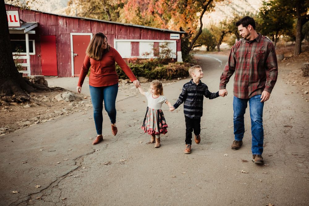 Family walking together on a path surrounded by autumn trees and red barn buildings in the background.