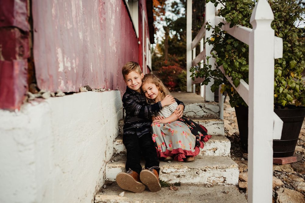 Two children sit together on white concrete steps outside a house with pink walls on a sunny day.