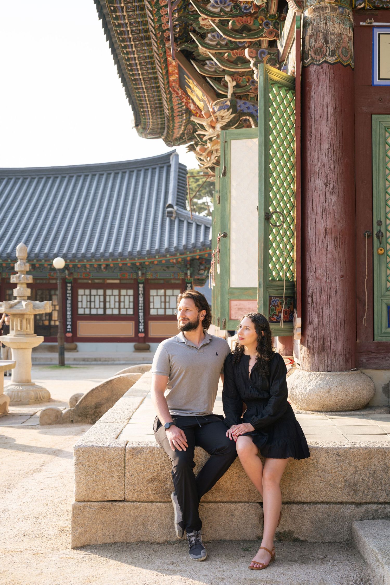 A couple sits together on traditional stone steps at a Korean temple with ornate wooden architecture in the background.