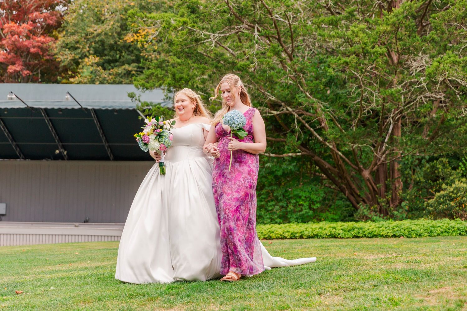 Two people in formal attire, one in a white gown and one in pink, walk together on a green lawn near trees.