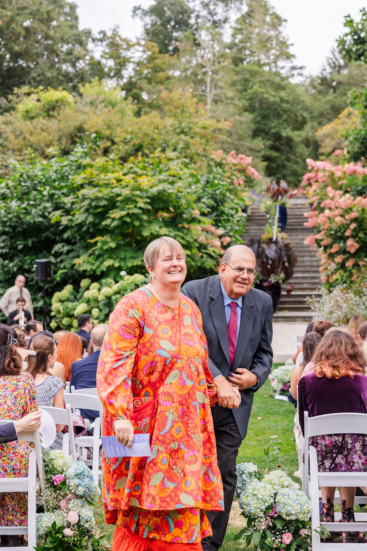 A joyful outdoor garden wedding ceremony with guests seated in white chairs among blooming hydrangeas and roses.