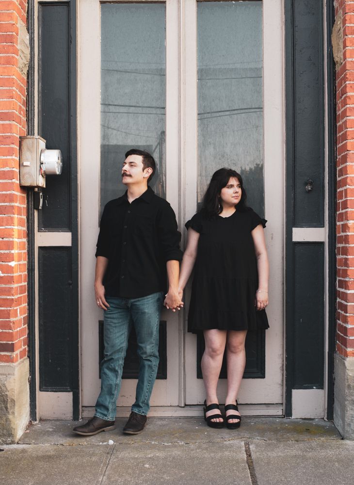 A couple in black outfits holding hands while standing in front of a glass door entrance with brick walls.