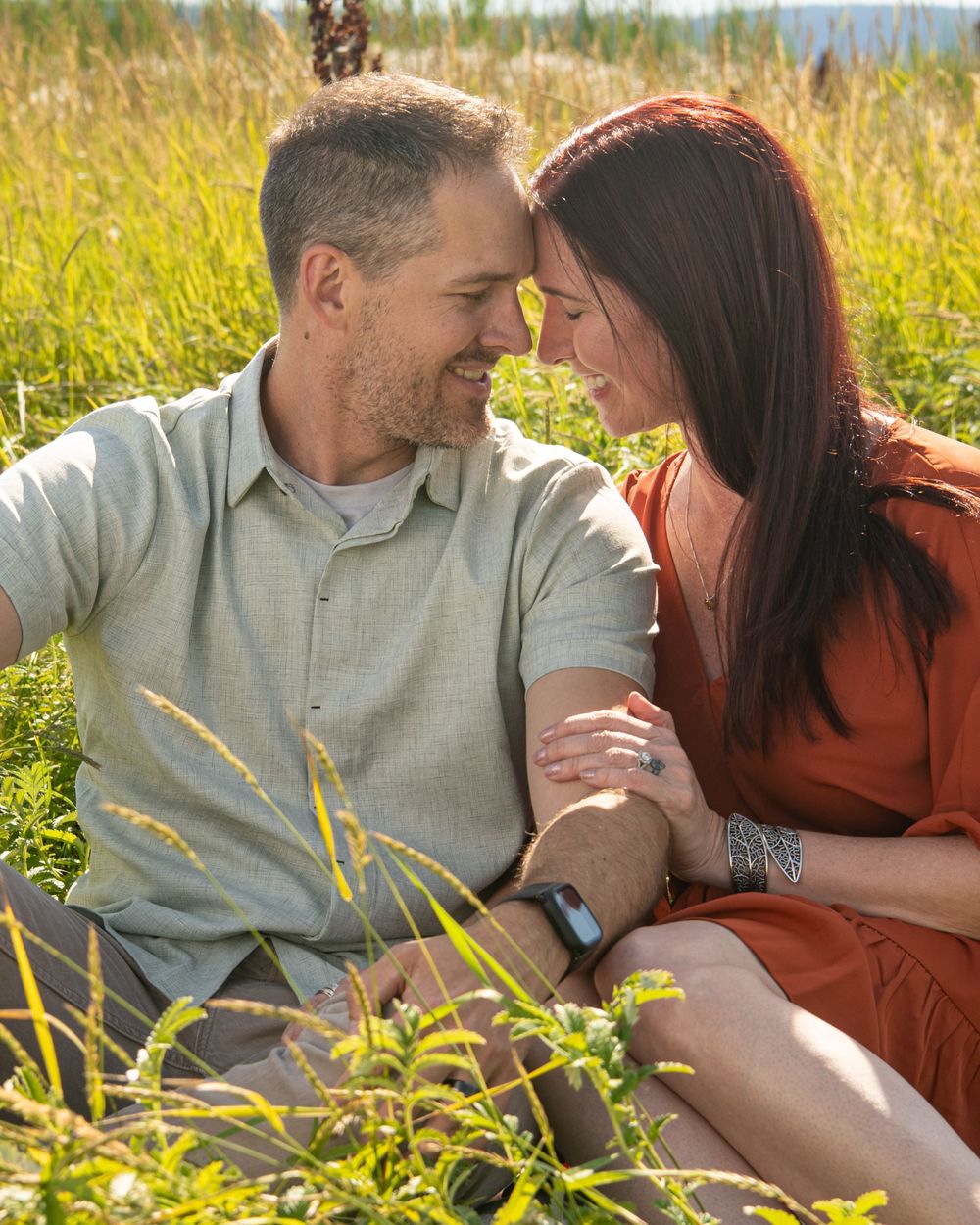A couple sits together in a sunlit meadow of tall grass and wildflowers sharing an intimate moment.