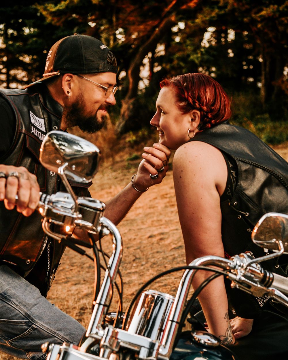 Two bikers share an intimate moment on their motorcycles at sunset in a wooded area.