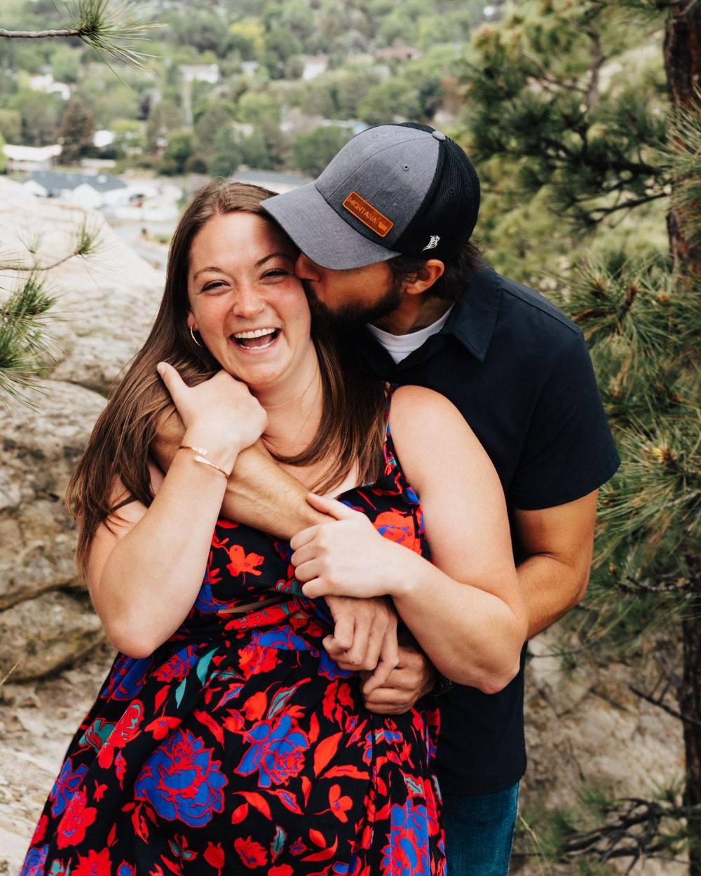 A couple shares a joyful moment together outdoors in a floral dress and baseball cap among pine trees on a mountainside.