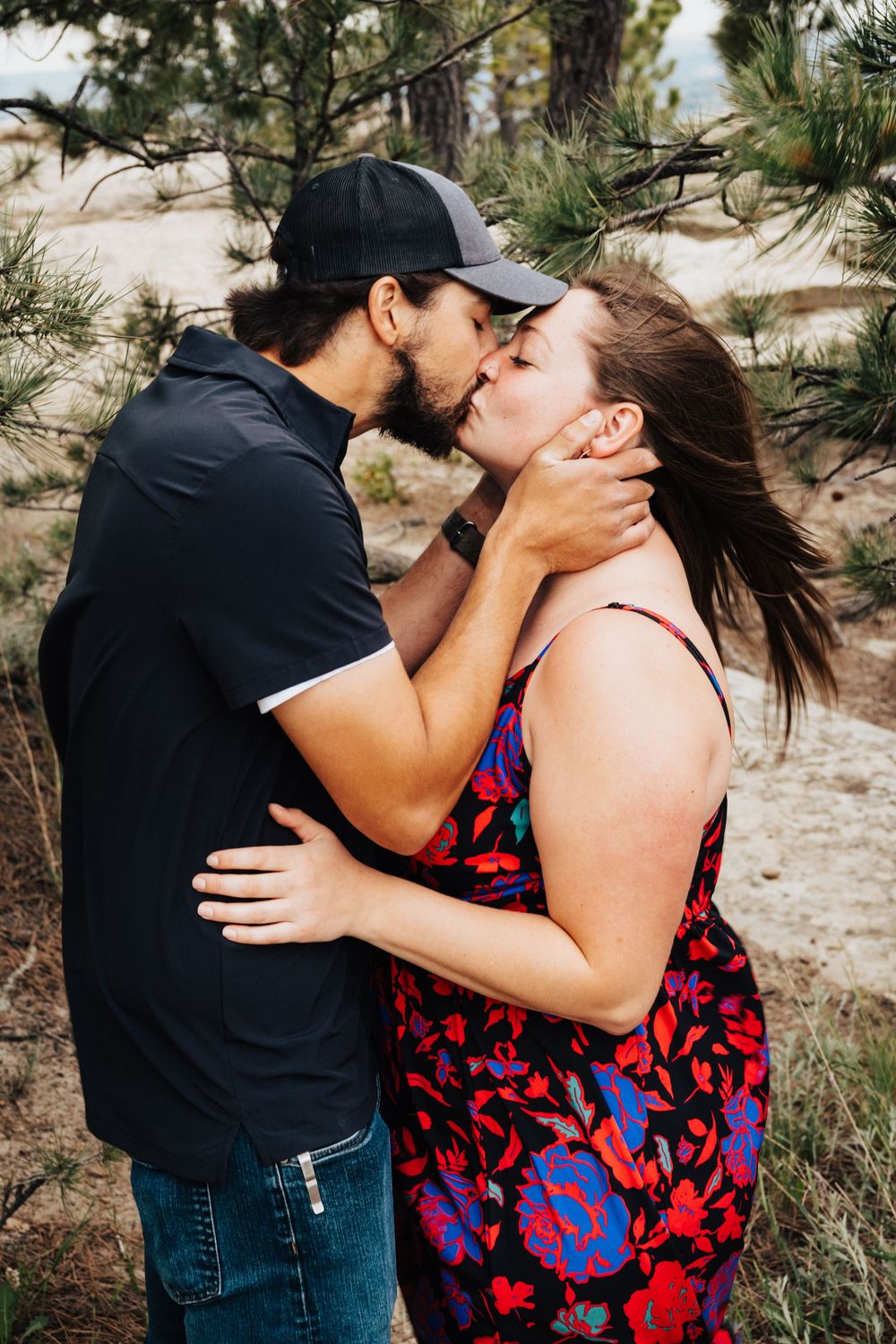 A couple shares a passionate kiss outdoors in nature, with the person wearing a red and blue floral dress.